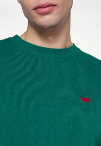 Gros plan d'une personne portant un pull côtelé vert avec une petite étiquette rouge sur la poitrine, montrant les lèvres et le cou.