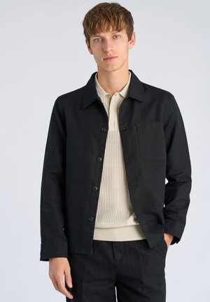 Schwarze Hemdjacket aus leichtem Material mit aufgespreiztem Kragen und einer einzelnen Brusttasche, darunter ein cremefarbenes Strickoberteil.