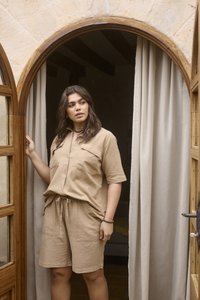 Femme en chemise beige boutonnée et short, debout dans une porte en bois cintrée avec des rideaux beiges, regardant sur le côté.