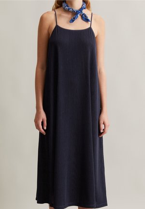 Femme portant une robe longue sans manches en maille côtelée bleu marine avec de fines bretelles et un foulard bleu à motifs autour du cou, sur un fond neutre.