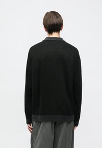 Crni pleteni džemper s dugim rukavima, visokim ovratnikom i teksturiranim završetkom, s kraćim stražnjim rubom i suptilnim sivim naglascima.