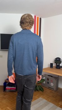 Plavi džemper dugih rukava od pletene tkanine, s opuštenim krojem. Sive hlače i drveni pod u modernom unutrašnjem okruženju.