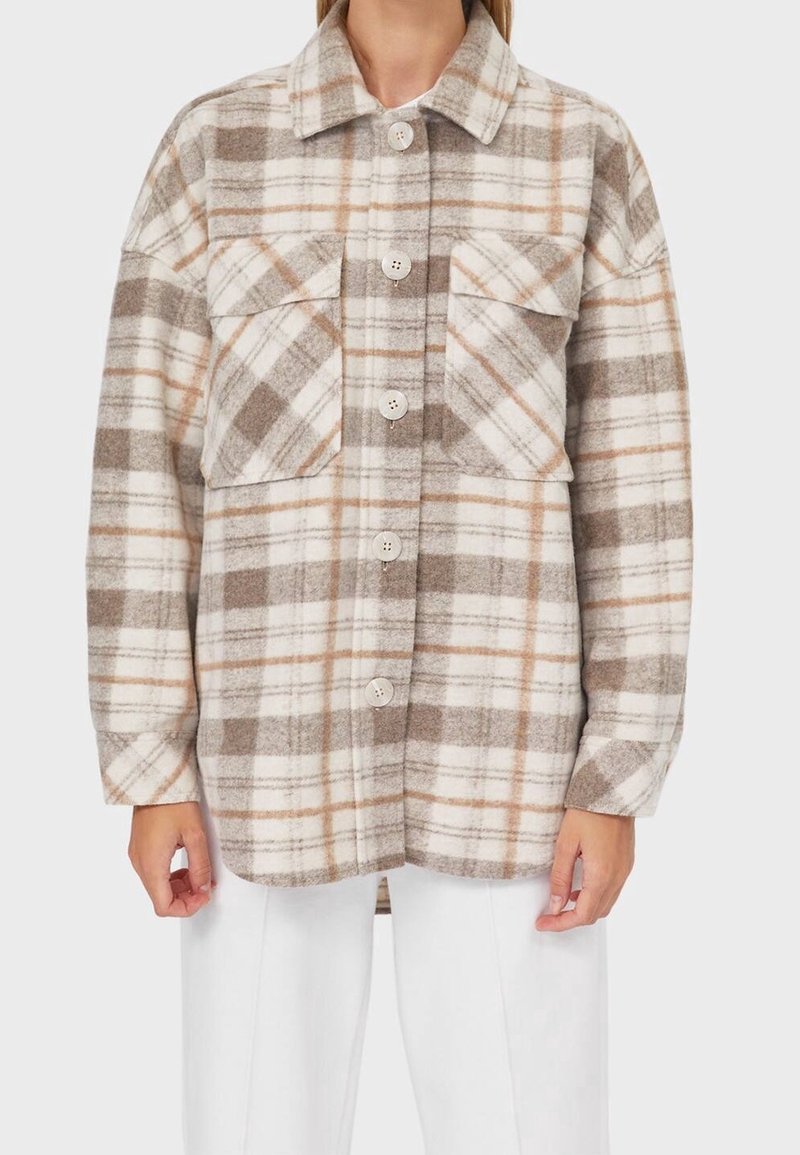 Femme portant une chemise à carreaux beiges, blanche et marron à boutons avec deux poches poitrine et un pantalon blanc, debout devant un fond uni.