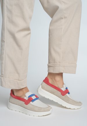 Sneakers in beige suede with red and blue accents. Rounded design, slip-on style, textured upper, and white sole. Corduroy pants shown.