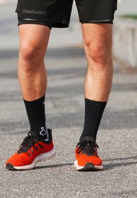 Scarpe sportive con una parte superiore in rete rossa e nera, suola texturizzata e lacci neri, abbinate a calzini neri alla caviglia con un design bianco.