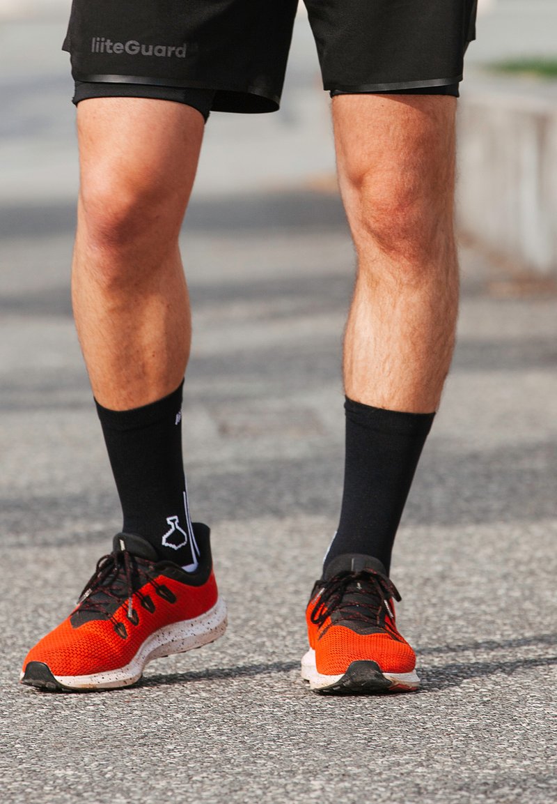 Scarpe sportive con una parte superiore in rete rossa e nera, suola texturizzata e lacci neri, abbinate a calzini neri alla caviglia con un design bianco.