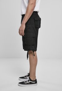 Person som bär svarta cargoshorts med flera fickor och svarta låga sneakers med vita snören, stående på sidan mot en neutral bakgrund.