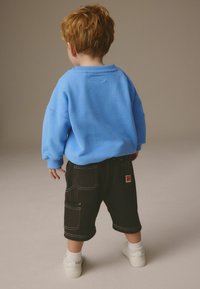Et ungt barn med rødt hår iført en blå sweatshirt, sorte shorts med lommer, hvide sokker og hvide sneakers, der står med ryggen til.