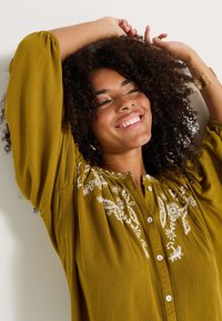 Blouse vert olive avec broderie florale blanche, fermeture à boutons à l'avant et manches longues bouffantes. Le tissu a une apparence texturée et froissée.