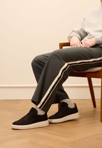 Schwarze Wildleder-Sneaker mit weißen Gummisohlen, kombiniert mit grauen Jogginghosen mit weißen Seitenstreifen, die auf einem Holzstuhl sitzen.