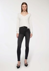 Maglione bianco a maniche lunghe con scollatura a V abbinato a jeans skinny scuri e tacchi a punta neri, che mettono in risalto un design aderente e texture lisce.