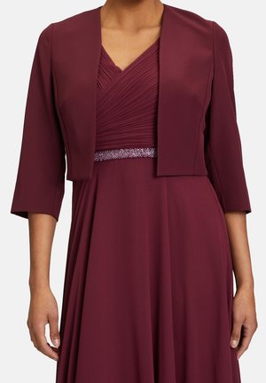 Femme portant une robe bordeaux foncé avec un corsage plissé en V, une veste courte assortie à manches trois-quarts, et une ceinture de taille ornée de perles.