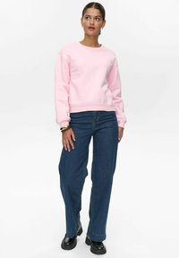 Rozā kokvilnas džemperis ar ribotām aprocēm, kombinēts ar zilām svītrām platām biksēm un melniem platforma zābakiem. Minimālas piederumu redzamas.