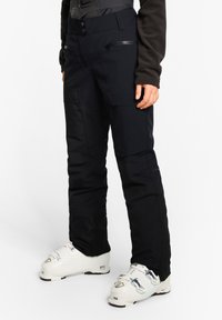 Schwarze Skihosen mit strukturierter Oberfläche, mit einem hochgeschnittenen Design, Reißverschlusstaschen und einer taillierten Silhouette, kombiniert mit weißen Skischuhen.