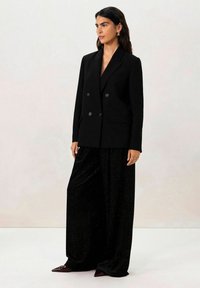 Blazer noir croisé en fils métalliques, associé à un pantalon large noir avec une texture scintillante. Chaussures pointues bordeaux.