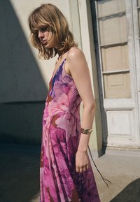 Dlouhé šaty bez rukávů z průsvitné látky s živým květinovým vzorem v odstínech růžové a fialové. Mají splývavou siluetu a jednoduchý design.