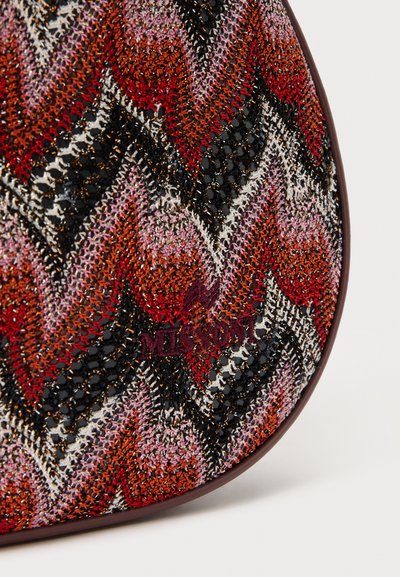 Τσάντα πολλών χρωμάτων με σχέδιο ζιγκ-ζαγκ σε κόκκινο, ροζ και μαύρο, φτιαγμένη από υφασμάτινο υλικό με υφή, με μια διακριτική ανάγλυφη λογότυπο μπροστά.