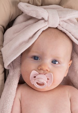 Vauva sinisin silmin makaa, yllään pehmeä vaaleanpunainen pyyhe, jossa on rusetti, ja käyttää vaaleanpunaista rusetin muotoista tuttia.