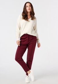 Pull en maille côtelée crème avec un décolleté en V, associé à un pantalon tailleur bourgogne avec coutures avant et des baskets blanches.