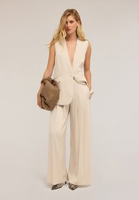 Beige mouwloze op maat gemaakte vest met diepe V-hals, gecombineerd met beige wijde broek. Houdt een bruine handtas vast en draagt puntige zwarte gestreepte hakken.