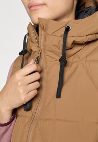 Gilet puffato marrone con colletto alto, caratterizzato da una chiusura con zip e cordini neri. Texture a trapunta e fodera interna a contrasto visibile.