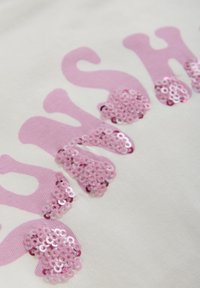 Witte stof met grote, zachte roze letters. De letters zijn voorzien van sprankelende roze pailletten, wat textuur en visuele interesse aan het ontwerp toevoegt.