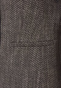 Tissu sombre texturé avec un motif en chevrons, présentant un léger éclat métallique et un détail de poche à rabat.