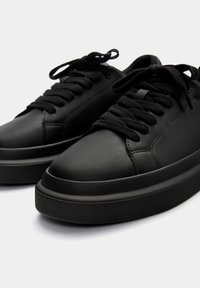 Fekete bőrből készült sneaker cipők lekerekített orral, lapos fűzőkkel és vastag, textúrázott talppal. Nincs látható márkázás vagy minta. Egyszerű, minimalista dizájn.