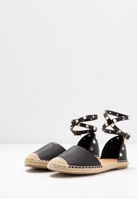 Espadrilles de cuero negro con punta afilada, suela de yute y correas en el tobillo con remaches. Parte superior texturizada y tapa de punta beige en contraste.