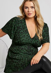 Top verde con stampa leopardata, scollatura a V profonda, maniche corte a sbuffo e silhouette fluida, abbinato a leggins di pelle nera.