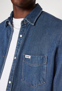 Chemise en denim bleu foncé avec un col pointu, des boutons pressions blancs et une poche affichant le logo Wrangler. Tissu texturé.