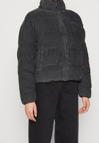 Svart kordfløjl puffjacka med hög krage, framdragkedja och texturerat vertikalt ribbat mönster. Tätt sittande silhuett med sidofickor.