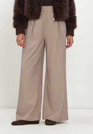 Woman wearing beige wide-leg pants with an elastic waist, brown textured jacket, and brown flat shoes standing against a plain background.
