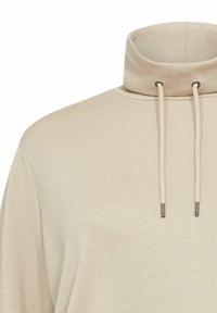 Beige pulóver felső magas gallérral és elülső húzózsinórral. Simaságot és puhaságot biztosító textúrával rendelkezik, finom varrási részletekkel a nyakvonalon.