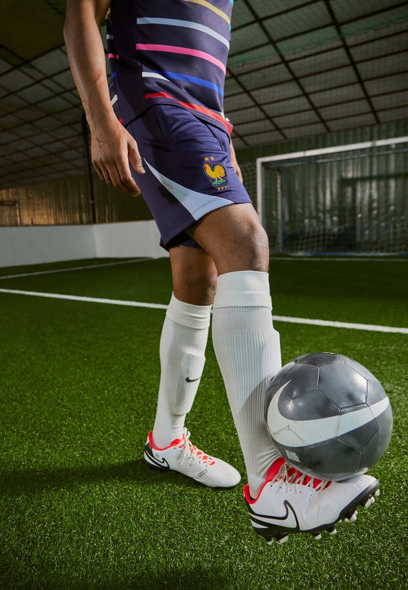 Voetballer in donkerblauw tenue met kleurrijke strepen, witte sokken en schoenen met rode accenten, die een zwart-grijze voetbal op het gras plaatst.