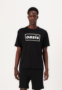 Juodas medvilnės marškinėliai su baltu stačiakampiu logotipu, kuriame parašyta "oasis". Atsipalaidavęs siluetas su trumpomis rankovėmis ir apvaliu iškirpimu.