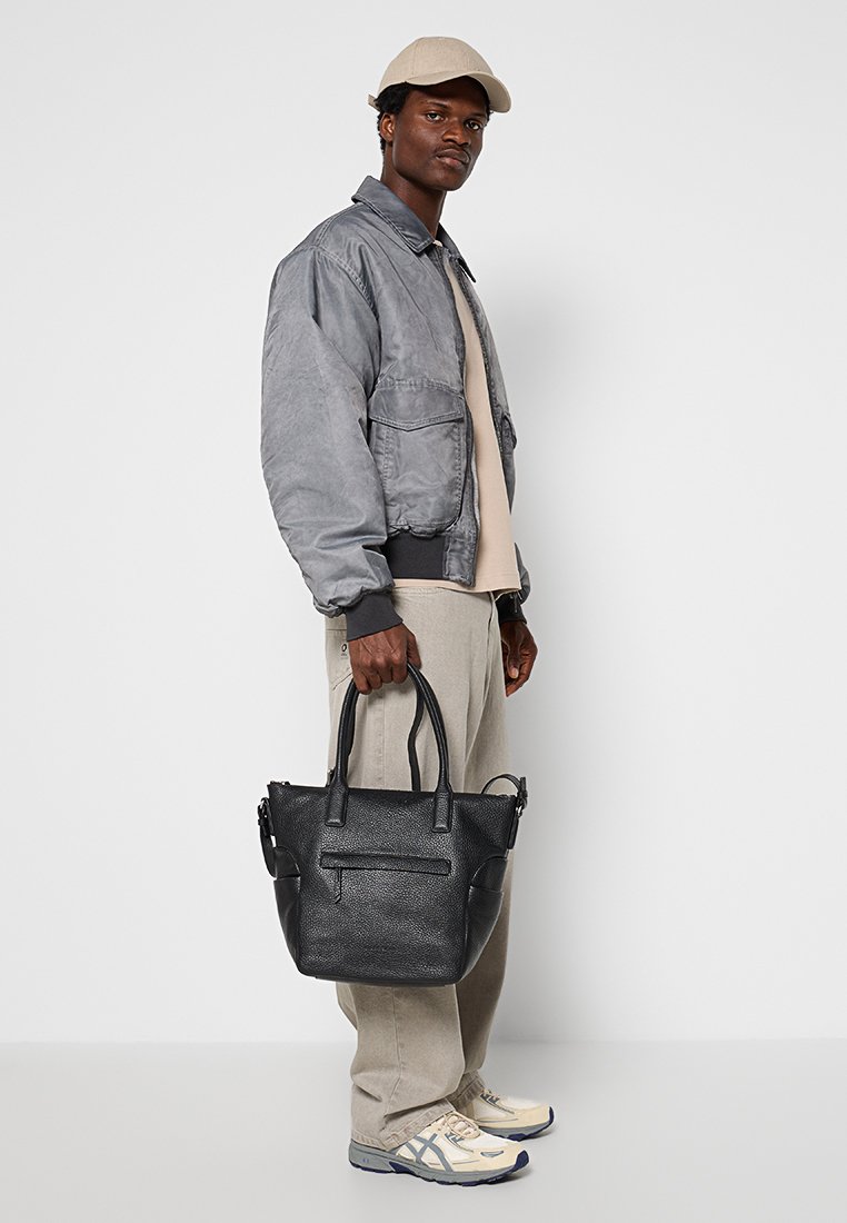 Schwarze Ledertasche mit doppelten Griffen, strukturiertem Äußeren, Fronttasche mit Reißverschluss und minimalistischen Design. Getragen von einer Person in legerer Kleidung.