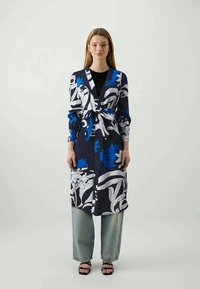 Μακριά κάρντιγκαν σε ναυτικό χρώμα με λουλουδάτα σχέδια σε μπλε και λευκό, δεμένο στη μέση. Φοριέται πάνω από μαύρη μπλούζα και συνδυάζεται με χαλαρές ανοιχτόχρωμες μπλε τζιν.