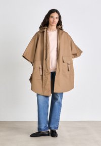 Imperméable marron surdimensionné avec capuche, équipé de grandes poches avant et de poignets ajustables. Porté par-dessus un haut rose clair et un jean bleu.