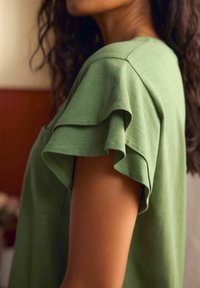 Zaļš īsroku tops ar volāniem piedurknēs, izgatavots no mīksta auduma. Tops ir vaļīga piegriezuma un ar smalku faktūru.