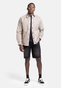 Veste beige clair à boutons avec deux poches poitrine, t-shirt noir en dessous, shorts en denim noir déchirés et baskets noires avec des accents blancs.