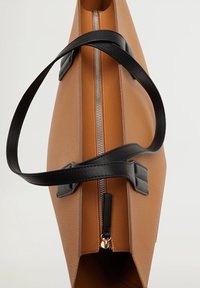 Brązowa torba tote z fakturowanej tkaniny, z czarnymi uchwytami i srebrnym zamkiem błyskawicznym. Ma szerokie otwarcie i płaskie dno dla stabilności.