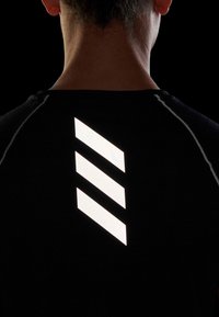 Zadní pohled na osobu v černém tričku s třemi reflexními bílými diagonálními pruhy na horní části zad, na černém pozadí.