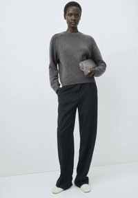Grauer Strickpullover mit Rundhalsausschnitt, zum schwarzen Nadelstreifenanzug kombiniert. Das Modell hält eine kleine graue Clutch und trägt weiße Slipper.