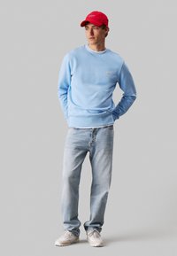 Ανοιχτό μπλε φούτερ με ριγωτές μανσέτες, λογότυπο Calvin Klein, σε συνδυασμό με ανοιχτό μπλε τζιν και μπεζ αθλητικά παπούτσια, συμπληρωμένο με κόκκινο καπέλο.