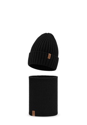 Homme portant un bonnet en maille noir et un chauffe-cou avec des étiquettes marron et une veste d'hiver grise sur un fond uni.