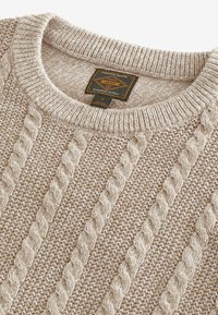 Beige Strickpullover mit geripptem Kragen und einem schwarzen Etikett mit der Aufschrift "Premium Grade 1982 Durable Goods" im Nacken.