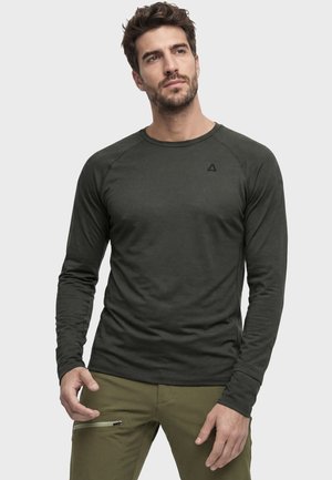 Mann mit kurzen dunklen Haaren, der ein eng anliegendes dunkelgrünes Langarmshirt und olivgrüne Hosen trägt, steht vor einem schlichten hellen Hintergrund.