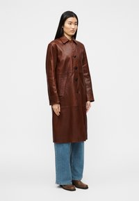 Καφέ δερμάτινο παλτό τραντσκότ με γιακά, κουμπιά μπροστά και πλαϊνές τσέπες, σε συνδυασμό με τζιν φαρδιάς γραμμής και καφέ υποδήματα.