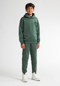 Felpa verde con un patch logo, abbinata a pantaloni jogger coordinati, caratterizzati da una texture liscia. Un paio di sneakers alte bianche completa l'outfit.